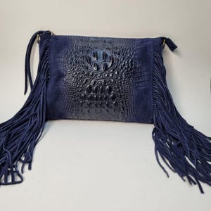 Puede incluir: Un bolso bandolera azul marino con un panel frontal con relieve de cocodrilo y laterales de ante. El bolso presenta largos flecos en los laterales y una correa para el hombro. Un accesorio elegante.