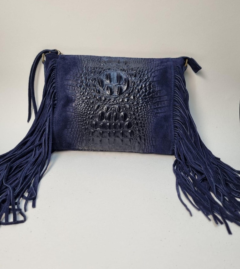 Puede incluir: Un bolso bandolera azul marino con un panel frontal con relieve de cocodrilo y laterales de ante. El bolso presenta largos flecos en los laterales y una correa para el hombro. Un accesorio elegante.