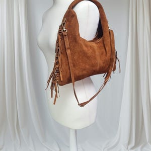 May include: A brown suede shoulder bag with a fringe detail. The bag has a curved shape and a long, adjustable strap. The bag is displayed on a white mannequin against a white backdrop.