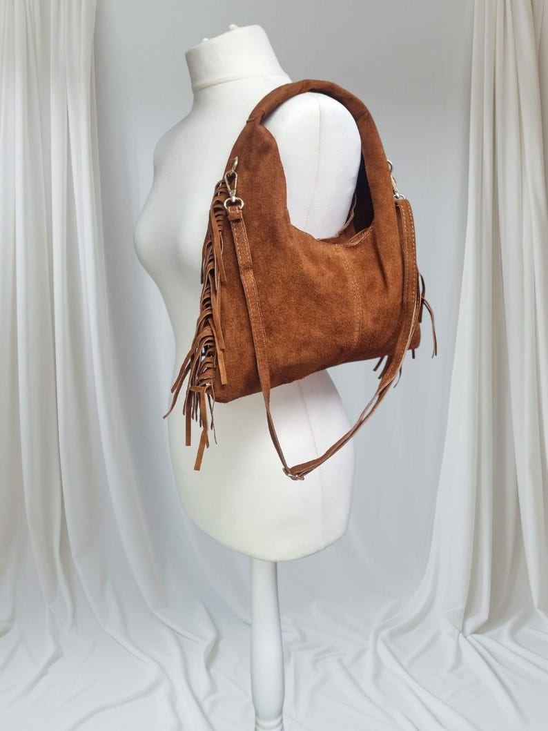 May include: A brown suede shoulder bag with a fringe detail. The bag has a curved shape and a long, adjustable strap. The bag is displayed on a white mannequin against a white backdrop.