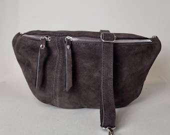 May include: A dark gray suede fanny pack with two zippered compartments and a silver-tone buckle. The adjustable strap and zipper pulls are made of the same suede material. The bag is designed to be worn around the waist or across the body.