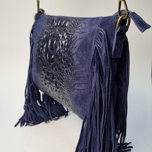 Puede incluir: Un bolso bandolera azul marino con un panel frontal texturizado con forma de cocodrilo. El bolso presenta largos flecos a los lados y una correa a juego. El bolso tiene un cierre color lat&oacute;n.