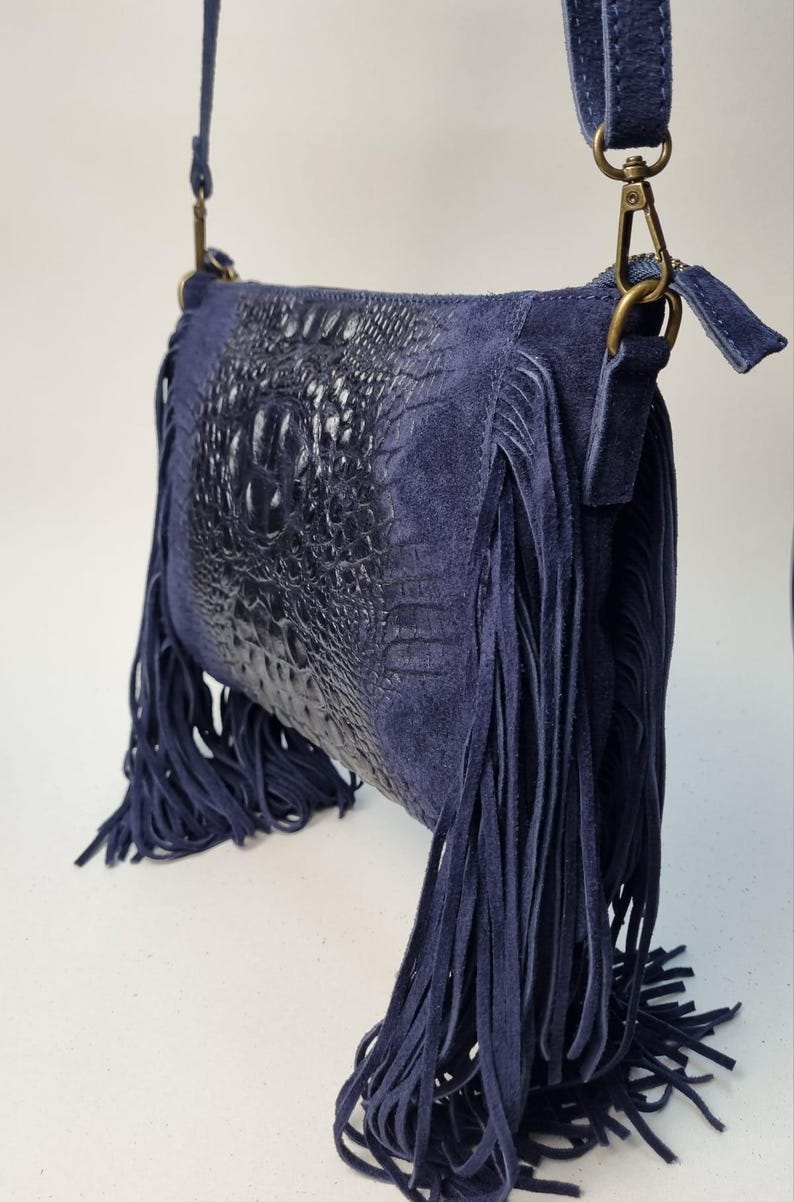 Puede incluir: Un bolso bandolera azul marino con un panel frontal texturizado con forma de cocodrilo. El bolso presenta largos flecos a los lados y una correa a juego. El bolso tiene un cierre color lat&oacute;n.