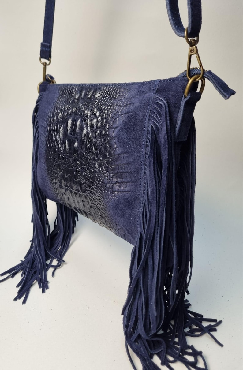 Puede incluir: Un bolso bandolera de ante azul marino con un panel central de cuero con relieve de cocodrilo. El bolso presenta largos flecos y una correa ajustable a juego. Los herrajes son de color lat&oacute;n.