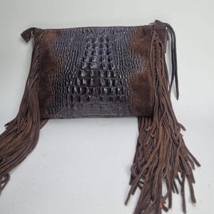 May include: A brown leather clutch bag with a crocodile-effect central panel and suede side panels. It has long, dangling fringe on both sides, giving it a bohemian style.