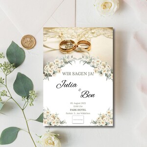 Könnte beinhalten: Eine weiße Hochzeitseinladung mit goldenen Ringen und floralen Akzenten. Der Text lautet "Wir sagen Ja!" gefolgt von den Namen "Julia & Ben", dem Datum "06. August 2023", der Uhrzeit "12:00 Uhr", dem Ort "PARK HOTEL" und der Adresse "Parkstr. 5, Am Wildchen".
