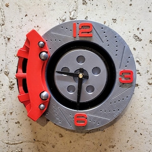 May include: A silver-colored clock designed to resemble a car brake rotor, with a red brake caliper on the left side. The clock face has red numerals at 12, 3, and 6 o'clock. The hands are black.