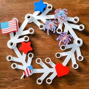May include: A white, snowflake-shaped wreath decorated with patriotic symbols. It features a red, white, and blue color scheme with a star, heart, American flag, and fireworks. The wreath is designed for festive holiday decor.