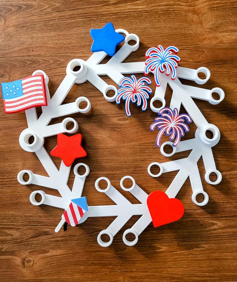 May include: A white, snowflake-shaped wreath decorated with patriotic symbols. It features a red, white, and blue color scheme with a star, heart, American flag, and fireworks. The wreath is designed for festive holiday decor.