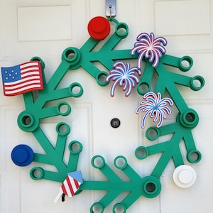 May include: A green wreath with red, white, and blue patriotic decorations. The wreath features an American flag, fireworks, and circular accents. It is hanging on a white door, perfect for celebrating Independence Day.