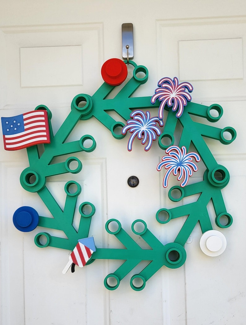 May include: A green wreath with red, white, and blue patriotic decorations. The wreath features an American flag, fireworks, and circular accents. It is hanging on a white door, perfect for celebrating Independence Day.