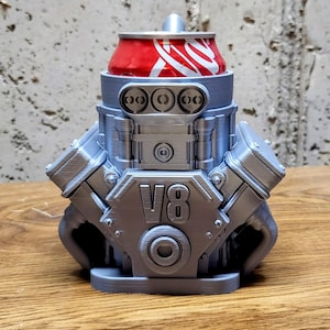 May include: A silver-colored, 3D-printed V8 engine-shaped can cooler holding a red Coca-Cola can. The cooler has detailed engine components and the text "V8" on the front. The cooler sits on a wooden surface.