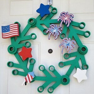 May include: A green wreath decorated with patriotic symbols. It features a small American flag, red and blue stars, and firework designs. The wreath is hanging on a white door.