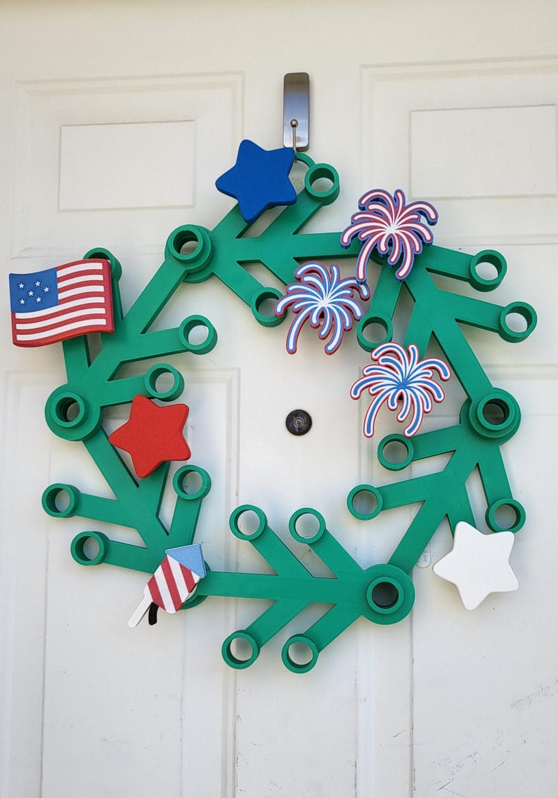 May include: A green wreath decorated with patriotic symbols. It features a small American flag, red and blue stars, and firework designs. The wreath is hanging on a white door.