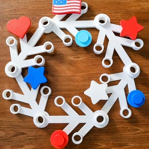 May include: A white, circular, modular toy with various shapes and colors. The design includes red hearts and stars, blue circles and stars, and a small American flag. The toy is set against a wooden background.