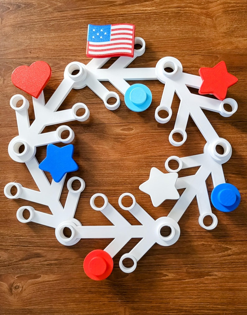 May include: A white, circular, modular toy with various shapes and colors. The design includes red hearts and stars, blue circles and stars, and a small American flag. The toy is set against a wooden background.