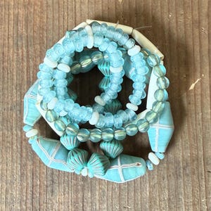 May include: A collection of handmade bracelets in shades of turquoise, white, and clear. The bracelets feature various bead shapes, including round, oval, and geometric designs. The beads are strung together with a visible cord, creating a layered effect.