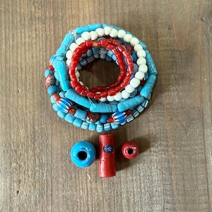 May include: A collection of colorful beads in shades of blue, red, and white. The beads are arranged in strands and individual pieces, showcasing various shapes and sizes. The beads are displayed on a wooden surface.