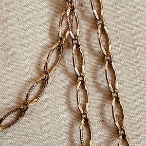 May include: Close-up of a gold-colored chain necklace with an oval link design. The chain is displayed against a neutral, textured background. The links have a textured pattern, adding detail to the jewelry.