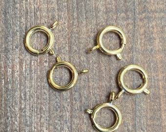 Op de afbeelding: Vijf goudkleurige metalen lobster-sluitingen, elk met een ronde ring en een veerbelaste sluiting. Deze sluitingen worden vaak gebruikt voor het maken van sieraden en knutselen.