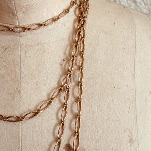May include: Gold-toned chain necklaces draped on a mannequin. The chains feature oval links with a textured pattern. The necklaces are layered, creating a cascading effect. The background is a neutral beige.