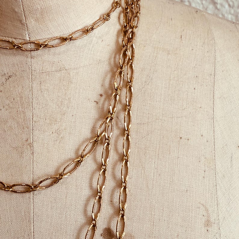May include: Gold-toned chain necklaces draped on a mannequin. The chains feature oval links with a textured pattern. The necklaces are layered, creating a cascading effect. The background is a neutral beige.