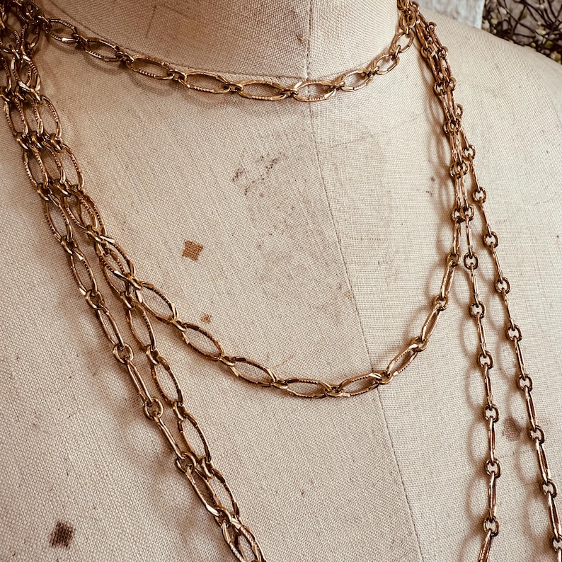 May include: Gold-toned necklaces with various chain link designs. The necklaces are draped on a mannequin, showcasing the different lengths and styles. The chains have a vintage aesthetic.