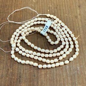 May include: A strand of off-white, irregularly shaped pearls. The pearls are strung together with a thin, white string. A small tag with the word "JAPAN" is attached to the strand. The pearls are arranged on a wooden surface.