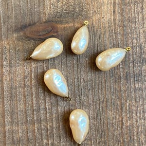 May include: Five off-white pearlescent teardrop-shaped beads with gold-toned metal loops. The beads are arranged on a wooden surface.
