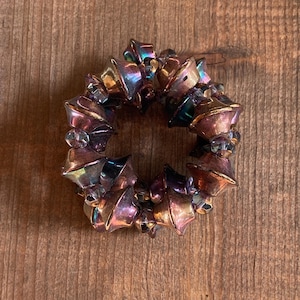 May include: A circular bracelet composed of iridescent, bell-shaped beads in shades of purple, gold, and teal. Clear, round beads are interspersed between the bell shapes. The bracelet is set against a wooden background.