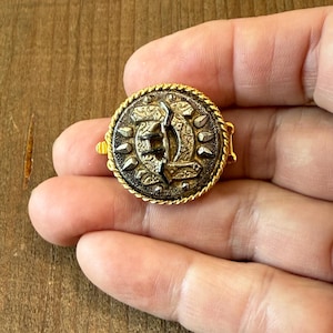 May include: A vintage-style button with a gold-tone border and intricate detailing. The button features a central design with a textured, metallic appearance. The button is round and has a small clasp on the back.