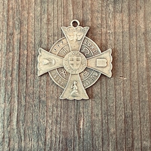 May include: A silver-toned cross pendant with religious symbols and text. The cross features a central circle with a smaller cross, surrounded by text and symbols. The words "ONE LORD" and "ONE FAITH" are visible. The pendant has a loop at the top for a chain.