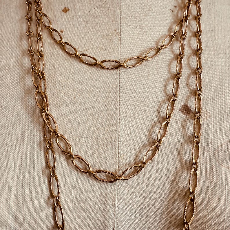 May include: Gold-toned chain necklaces with oval links. The necklaces are layered, with one longer chain and a shorter one. The chains have a textured, vintage appearance, and are displayed against a neutral background.