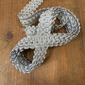 May include: Silver decorative trim with a looped design. The trim is made of a shiny, metallic material and is arranged in a loose, coiled pattern on a wooden surface. This trim is suitable for various craft and sewing projects.
