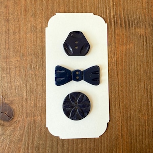May include: Three navy blue buttons in different shapes: a geometric shape with two holes, a bow tie, and a round button with a floral design. The buttons are arranged on a white card against a wooden background.