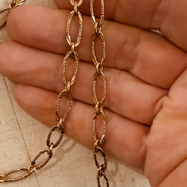 May include: Close-up of a gold-colored chain necklace with oval-shaped links. The links have a textured, detailed design. The necklace is displayed against a neutral background, highlighting the jewelry's color and texture.