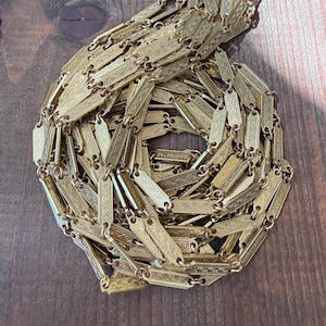 May include: A gold-toned metal chain necklace composed of numerous elongated rectangular links, each featuring a repeating etched cross-hatch pattern. The chain is coiled into a circular shape, showcasing the intricate texture of the metal links.