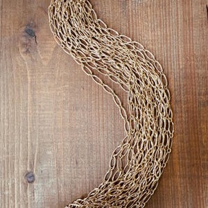 May include: A gold-colored necklace with a woven chain design. The necklace is draped on a wooden surface, showcasing its intricate pattern and texture. The warm tones of the gold contrast with the wood grain.