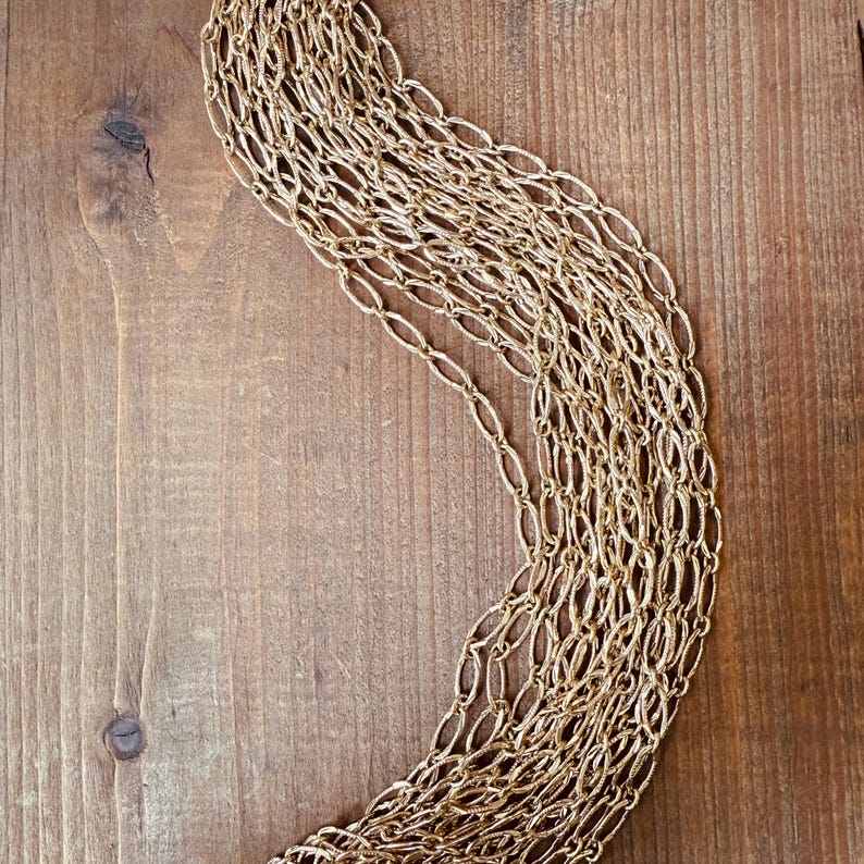 May include: A gold-colored necklace with a woven chain design. The necklace is draped on a wooden surface, showcasing its intricate pattern and texture. The warm tones of the gold contrast with the wood grain.