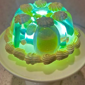 Op de afbeelding: Een gloeiend, groen en blauw gelatine dessert met een tulbandvorm, versierd met crème-kleurig glazuur. Het dessert staat op een witte taartstandaard, van binnenuit verlicht.