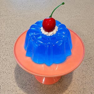 Puede incluir: Un postre de gelatina azul brillante moldeado en forma estriada, cubierto con una porción de crema blanca y una cereza roja con tallo verde, todo sobre un soporte para pasteles de color rosa claro con una base circular lisa.
