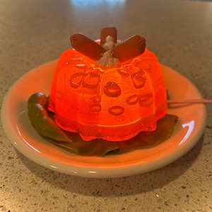 May include: A bright orange gelatin mold with a textured surface, topped with three small brown hot dogs. The mold is sitting on a white plate with a green leaf.