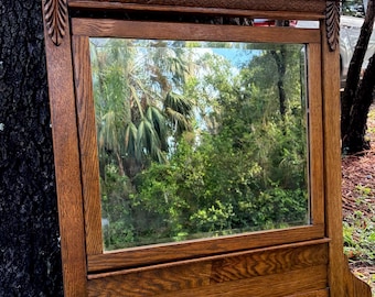 May include: A rectangular wooden mirror with a medium brown finish. The frame features a carved crest at the top, a cylindrical horizontal moulding, and decorative leaf-like carvings on the side pillars. The mirror glass reflects trees.