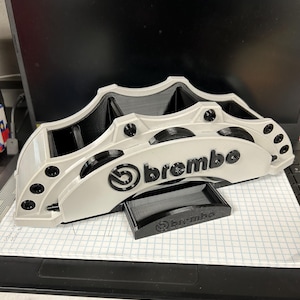 May include: A white and black Brembo brake caliper model. The model features the Brembo logo and is made of plastic. The caliper is resting on a piece of graph paper. A smaller black piece with the Brembo logo is in front of the caliper.