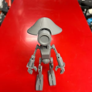 May include: A gray 3D printed robot toy with a round head and a wide, flat, gray hat. The robot has articulated arms and legs.