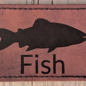 Könnte beinhalten: Ein rechteckiger, rotbrauner Lederaufnäher mit einer schwarzen Fischsilhouette und dem Wort "Fish" in fetter, serifenloser Schrift. Der Aufnäher hat einen genähten Rand.