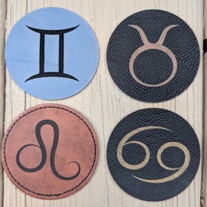 May include: Four round coasters featuring zodiac symbols: Gemini (blue), Taurus (dark blue), Leo (red-brown), and Cancer (dark blue). Each coaster has a stitched edge and a unique symbol design.