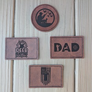 May include: Four brown leather patches with black text and designs. The patches feature a deer hunting scene, the word "DAD" with a deer and a target, a deer with the text "Deer Hunting Season", and a deer with a flag.