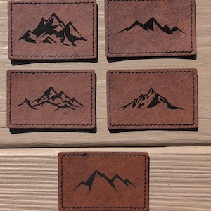 Può includere: Cinque toppe in pelle marrone con silhouette di montagne nere. Le toppe hanno un bordo cucito e sono disposte su due file su una superficie di legno.