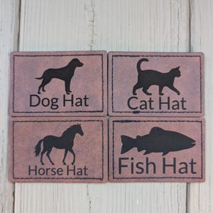 Könnte beinhalten: Vier rechteckige Aufnäher mit schwarzen Silhouetten eines Hundes, einer Katze, eines Pferdes und eines Fisches. Jeder Aufnäher trägt die Wörter "Dog Hat", "Cat Hat", "Horse Hat" und "Fish Hat" in Schwarz gedruckt. Die Aufnäher sind hellbraun.